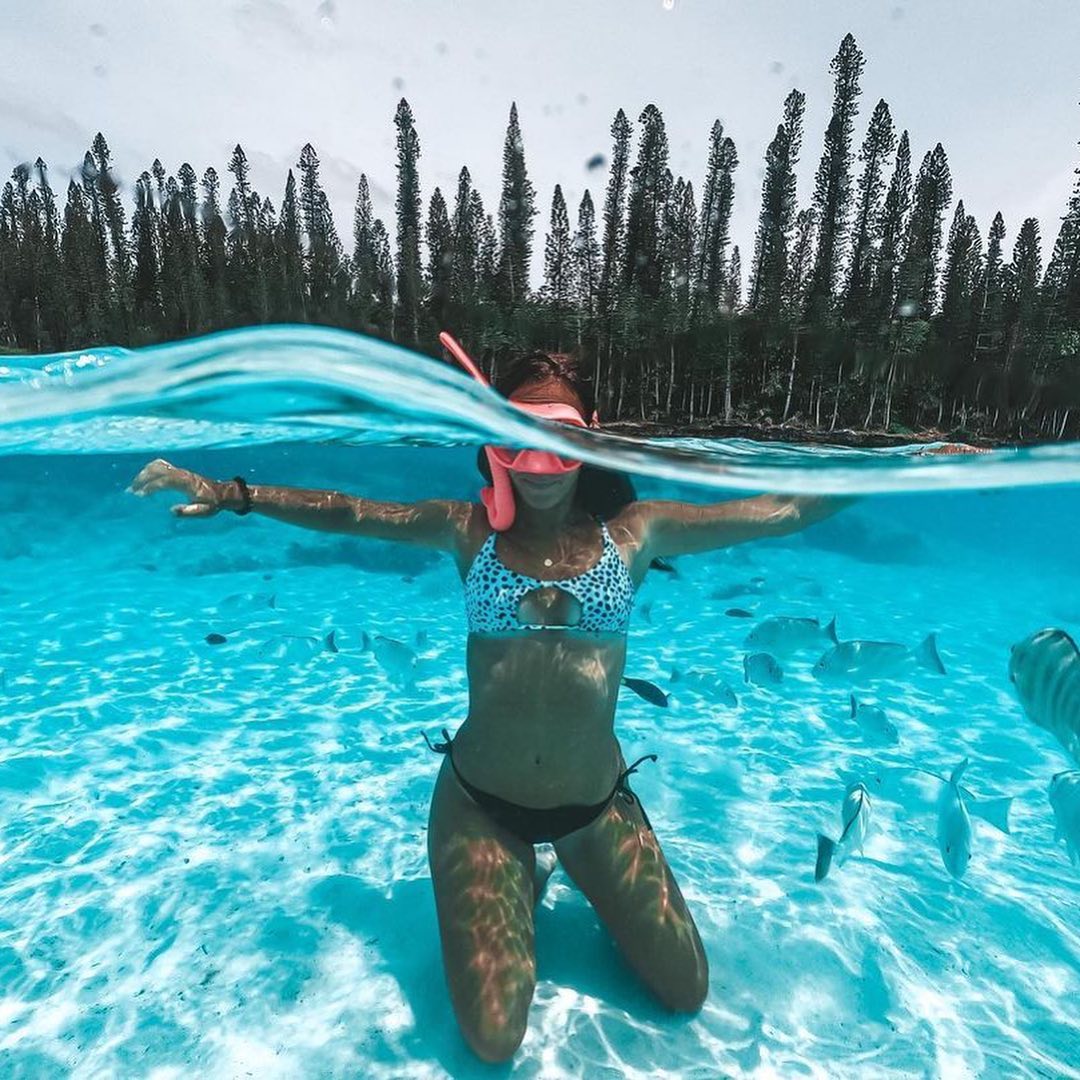 Une femme est à genoux dans une eau de mer transparente. Elle est en maillot de bain et porte un masque de plongée sous-marine Maxlux S Beuchat Rose et le tuba Spy associé de la même couleur. En arrière plan on voit la plage et des arbres. 